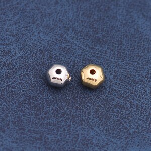 May include: Two small, faceted beads. One bead is silver-colored, and the other is gold-colored. The beads have a geometric shape with multiple sides and a hole through the center. The beads are displayed on a blue surface.
