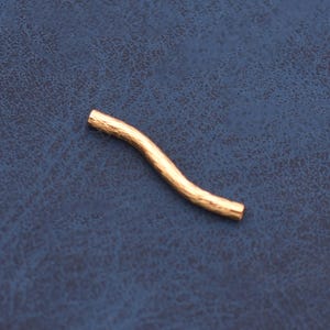 May include: A single, small, curved gold-toned metal tube bead with a textured, organic surface finish. The bead features a gentle S-shaped wave, designed for jewelry making, and is presented as a solitary piece against a dark blue background.