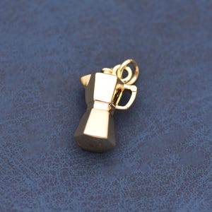 May include: A small, polished gold-toned metal charm shaped like a traditional stovetop moka pot coffee maker. The charm features a faceted, geometric body with a small handle on the side and a circular jump ring attached to the top for hanging.