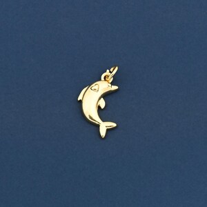 May include: A gold-colored dolphin charm with a small loop at the top for attaching to a necklace or bracelet. The dolphin has a heart-shaped marking on its side and is set against a dark blue background.
