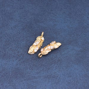 Op de afbeelding: Twee goudkleurige veervormige bedels met kleine, heldere edelstenen. De bedels hebben een getextureerd veermotief en een lus aan de bovenkant om aan sieraden te bevestigen. De achtergrond is effen donkerblauw.