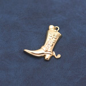 May include: A gold-toned metal charm shaped like a cowboy boot, featuring an embossed floral pattern on the shaft, a defined heel, and a small circular jump ring attached at the top for hanging, with a polished, reflective surface finish.