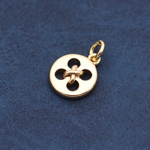 May include: A gold-colored button charm with a four-petal design and a loop for attaching to a necklace or bracelet. The charm has a smooth, polished finish and is set against a dark blue background.