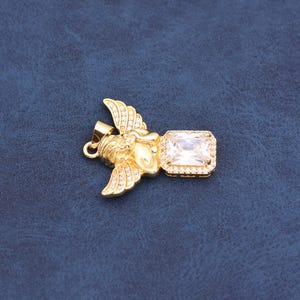 May include: A gold-coloured pendant depicting an angel with outstretched wings and a rectangular, clear gemstone. The angel's wings and the gemstone's frame are adorned with small, sparkling stones. The pendant is set against a dark blue background.