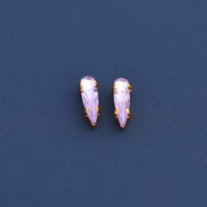 May include: A pair of light purple teardrop-shaped earrings with gold-colored metal accents. The earrings are set against a dark blue background, highlighting their delicate design and color.