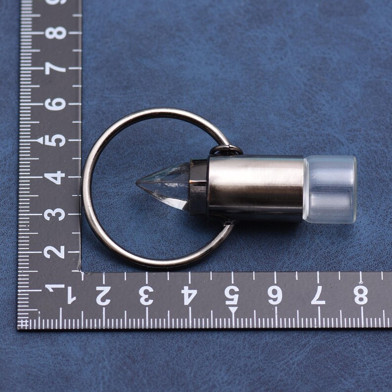 May include: A silver-colored keychain with a clear, faceted crystal point and a clear, rectangular end. The keychain is attached to a silver ring. The object is placed on a blue surface next to a ruler.
