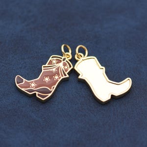May include: Two gold-colored cowboy boot charms. One charm is brown with white star details and a bow design. The other charm is a solid gold color. Both charms have a loop at the top for attaching to a necklace or bracelet.