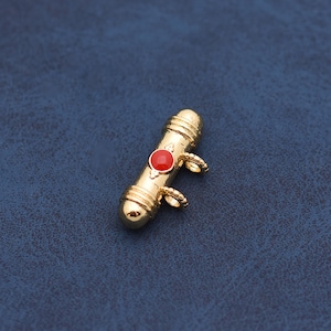 May include: A gold-colored pendant with a cylindrical shape, featuring a red circular stone in the center. The pendant has decorative bands and small loops on each side. The background is a dark blue.