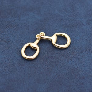 May include: A gold-colored equestrian-style snaffle bit charm. The charm features two rounded rings connected by a central bar with a looped design. The metal has a polished, reflective surface, and the background is a textured, dark blue.