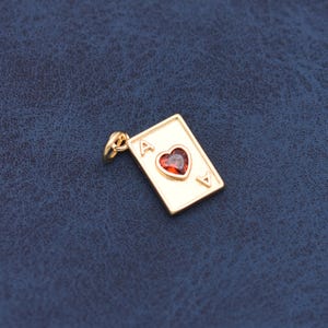 May include: A gold-toned metal charm shaped like a rectangular playing card. The charm features the letter A in the top left and bottom right corners, with a central heart-shaped red gemstone set in the middle of the card face.