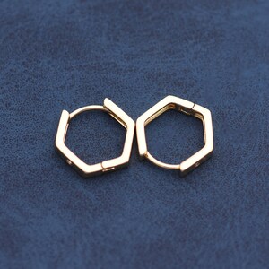 May include: A pair of gold-colored hexagonal hoop earrings. The earrings have a geometric design and a simple clasp closure. The earrings are displayed on a dark blue surface.