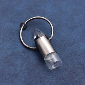 May include: A silver-colored keychain with a clear, bullet-shaped pendant. The pendant has a pointed crystal top and a cylindrical base. The keychain ring is circular and made of the same silver material. The background is a textured blue surface.