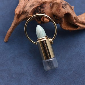 May include: A gold-colored essential oil bottle with a light blue gemstone top and a clear glass base. The bottle is attached to a gold ring. The background features a piece of driftwood on a blue surface.