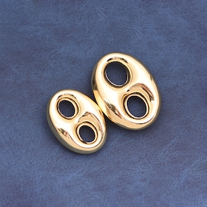 May include: Two gold-colored, oval-shaped jewelry components with two circular holes each. The components are displayed on a textured, dark blue surface. The shiny, metallic pieces reflect light, highlighting their smooth, polished finish.