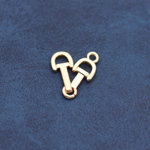 May include: Two gold-colored metal clasps, each with a D-shaped loop and a rectangular bar. One clasp has a small loop at the top. The clasps are arranged together on a dark blue surface. These clasps are likely used for jewelry or other craft projects.