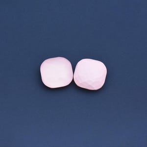 Könnte beinhalten: Zwei hellrosa, geometrisch geformte Objekte mit facettierter Oberfläche vor einem dunkelblauen Hintergrund. Die Objekte haben eine abgerundete quadratische Form und scheinen aus einem festen Material zu bestehen.