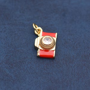 May include: A gold-colored charm in the shape of a vintage camera. The camera body is red with a gold-colored lens and accents. The lens features a clear, round gemstone. The charm has a small loop for attaching to a necklace or bracelet.
