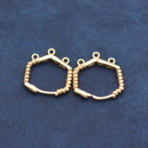 May include: Two gold-colored, geometric-shaped earring components. Each component has a hexagonal top with a small loop and a curved, beaded bottom. The earrings are displayed on a dark blue surface.