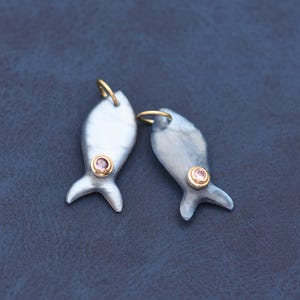 May include: Two fish-shaped pendants crafted from iridescent grey shell material. Each pendant features a small, round, light pink gemstone set in a gold-toned bezel. A gold-toned jump ring is attached to the top of each fish-shaped charm.