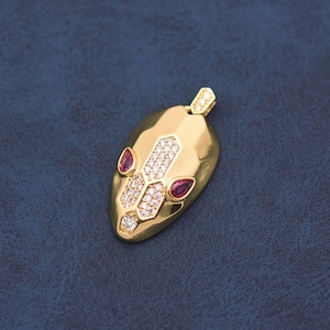 May include: A gold-toned pendant shaped like a snake head, featuring a faceted surface, a central inlay of clear pavé-set cubic zirconia stones, and two pear-shaped red stones serving as eyes, with a small bail at the top for attachment.