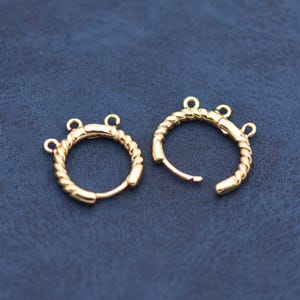 Op de afbeelding: Twee goudkleurige oorringen met een gedraaid touwontwerp. Elke oorring heeft drie kleine lussen voor bevestiging. De oorbellen worden gepresenteerd op een donkerblauwe ondergrond. Diameter ongeveer 2,5 cm.