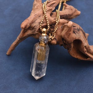 Puede incluir: Un collar con colgante de cristal transparente con una cadena dorada. El colgante tiene forma de prisma rectangular con un interior dorado. El collar se exhibe sobre un trozo de madera flotante sobre un fondo azul.