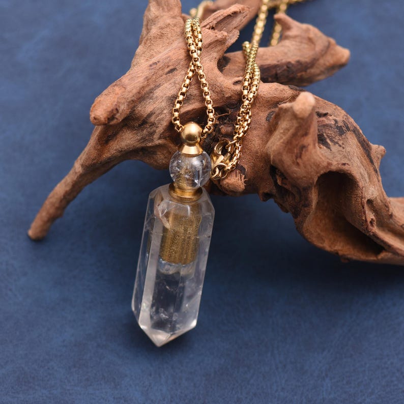 Puede incluir: Un collar con colgante de cristal transparente con una cadena dorada. El colgante tiene forma de prisma rectangular con un interior dorado. El collar se exhibe sobre un trozo de madera flotante sobre un fondo azul.
