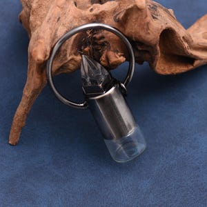 May include: A silver-colored bullet-shaped pendant with a clear glass bottom, attached to a metal ring. The pendant has a faceted crystal top. The background features a piece of driftwood on a blue surface.