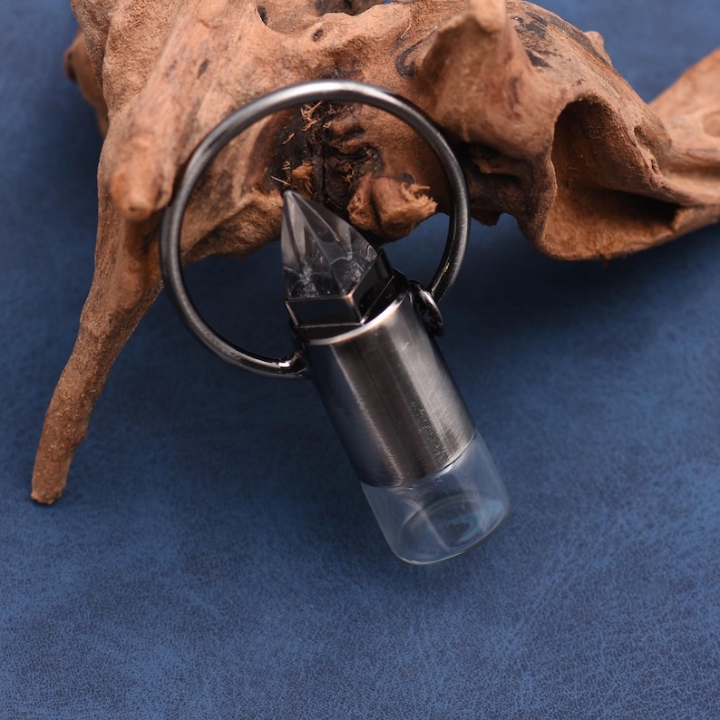 May include: A silver-colored bullet-shaped pendant with a clear glass bottom, attached to a metal ring. The pendant has a faceted crystal top. The background features a piece of driftwood on a blue surface.