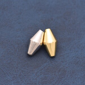Könnte beinhalten: Zwei bikonische Perlen, eine mit poliertem silberfarbenem Finish und die andere mit poliertem goldfarbenem Finish. Jede Perle weist ein facettiertes, konisches Design auf, das sich nach oben und unten verjüngt und ein symmetrisches Profil bildet.