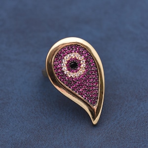Könnte beinhalten: Goldfarbener Ring mit tropfenförmiger Fassung. Die Fassung ist mit kleinen, runden, magentafarbenen Steinen gefüllt, mit einem zentralen schwarzen Stein, der von weißen Steinen umgeben ist. Der Ring liegt auf einer blauen Oberfläche.