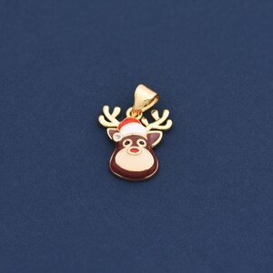 May include: A gold-colored pendant in the shape of a reindeer with a red and white Santa hat. The reindeer has brown antlers, a brown nose, and a small crystal. The pendant is on a dark blue background.
