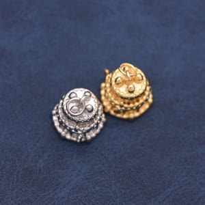 May include: Two small, three-tiered cake charms, one in a silver tone and one in a gold tone. Each charm features a circular jump ring attached to the top, with textured detailing representing layers and decorative dots on the surface of the cake.