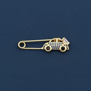 Könnte beinhalten: Eine goldfarbene Sicherheitsnadel mit einem charmanten Autoanhänger. Der Autoanhänger ist mit kleinen blauen und schwarzen Steinen sowie einem rechteckigen weißen Stein verziert. Die Nadel befindet sich vor einem dunkelblauen Hintergrund.