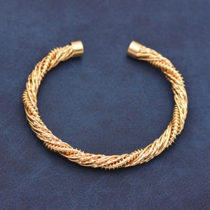 May include: A gold-toned cuff bracelet featuring a twisted, multi-strand wire design. The bracelet has a circular shape with an open gap at the top, finished with smooth, cylindrical end caps on both sides of the opening, creating a textured look.