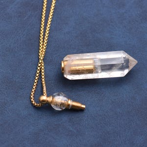 Puede incluir: Un collar de cadena dorada con dos colgantes de cristal. Un colgante es un cristal transparente y alargado con detalles met&aacute;licos dorados. El otro colgante es un peque&ntilde;o orbe transparente con detalles met&aacute;licos dorados.