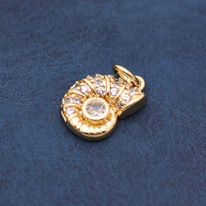 May include: A gold-colored pendant in the shape of a nautilus shell, encrusted with small, clear gemstones. The pendant has a loop at the top for attaching to a chain. The background is a dark blue.