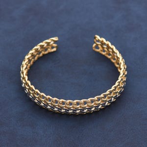 May include: A gold-colored chain bracelet with a textured, woven design. The open cuff bracelet is displayed on a dark blue surface. The bracelet is made of multiple rows of interlocking chain links.