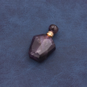 May include: A faceted, amethyst-colored perfume bottle with a gold-colored band around the neck and a faceted stopper. The bottle has a geometric shape and sits on a blue surface.