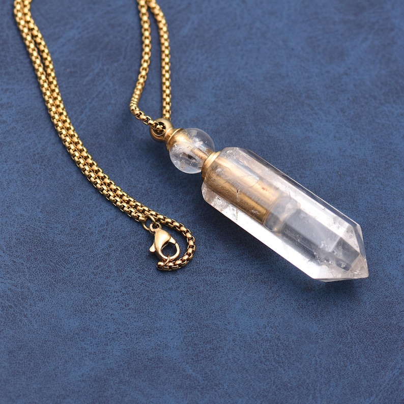 Puede incluir: Un collar de cadena dorada con un colgante de cristal transparente. El colgante es un cristal transparente y alargado con una carcasa de metal dorado. El collar est&aacute; sobre una superficie azul.