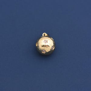 May include: A gold-colored soccer ball pendant with a small loop at the top for a chain. The ball features a classic pentagon and hexagon design, with small, sparkling accents. The pendant is set against a dark blue background.