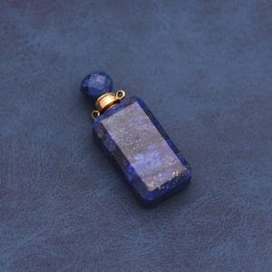 Może przedstawiać: Prostokątny, ciemnoniebieski flakon perfum z lapis lazuli z fasetowanym, kulistym korkiem. Butelka ma złote metalowe akcenty i małą pętlę na łańcuszek. Kamień ma złote plamki.