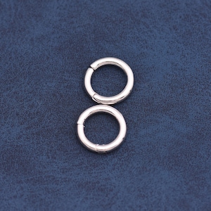 May include: Two silver-colored, circular clasps with a hinged opening. The clasps are shown against a textured, dark blue background. The clasps are likely used for jewelry or other small accessories.