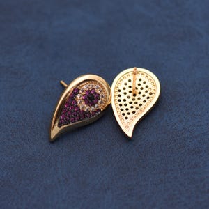 May include: Gold-coloured teardrop earrings with a stud post. The earrings feature a design of small, round, purple and white gemstones. The back of one earring is visible, showing a perforated pattern.