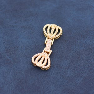 May include: A gold-colored jewelry clasp with a crown design. The clasp features two crown-shaped elements connected by a rectangular bar, all encrusted with small, sparkling stones. The background is a textured, dark blue surface.