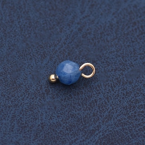 Può includere: Una piccola perla blu sfaccettata con un anello color oro e una perla d'oro più piccola. La perla blu è il punto focale, con gli elementi dorati che aggiungono contrasto. Il componente del gioiello è posto su uno sfondo blu scuro.