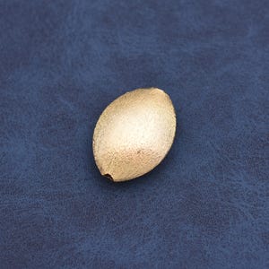 May include: A gold-colored, oval-shaped bead with a textured surface. The bead has a small hole at one end, suggesting it is for stringing or attaching to other items. The bead is set against a dark blue background.