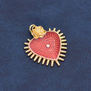 May include: A gold-toned pendant in the shape of a heart with a red enamel finish. The heart features a central eye design with a small, clear gem and is topped with a gold rose. The pendant has a sunburst design around the heart.