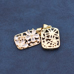 Op de afbeelding: Twee goudkleurige rechthoekige hangers. Eén hanger heeft een bloemenontwerp met heldere kristallen, de andere heeft een uitgesneden bloemenpatroon. Beide hangers hebben een lus voor een ketting.