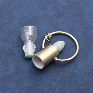 May include: A clear glass roller bottle and a gold-colored metal rollerball with a light green stone tip. The rollerball is attached to a gold-colored ring. The items are on a dark blue surface.